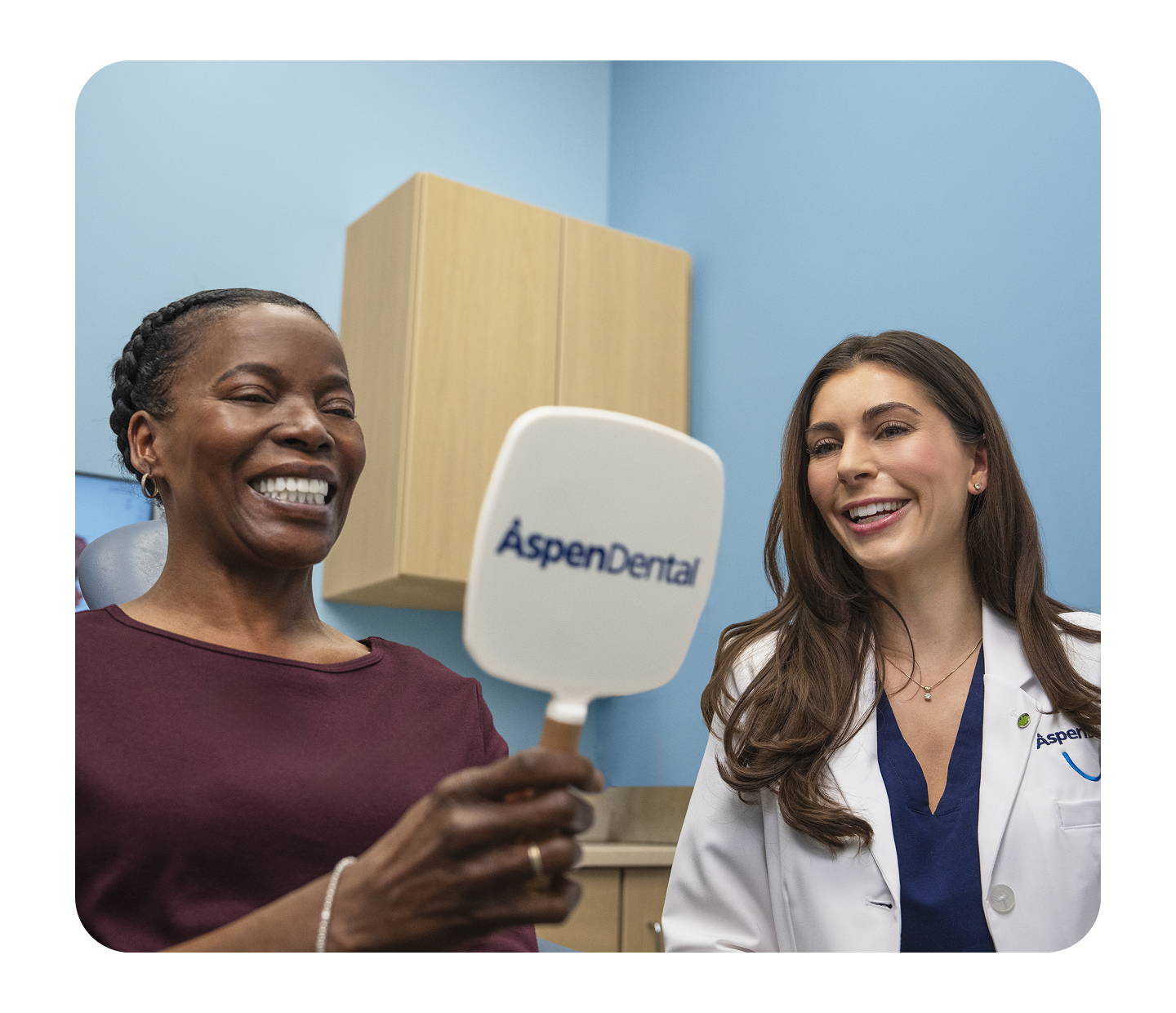 A smiling patient looks into an Aspen Dental mirror while a dentist in a white coat stands beside her, smiling during a dental appointment.