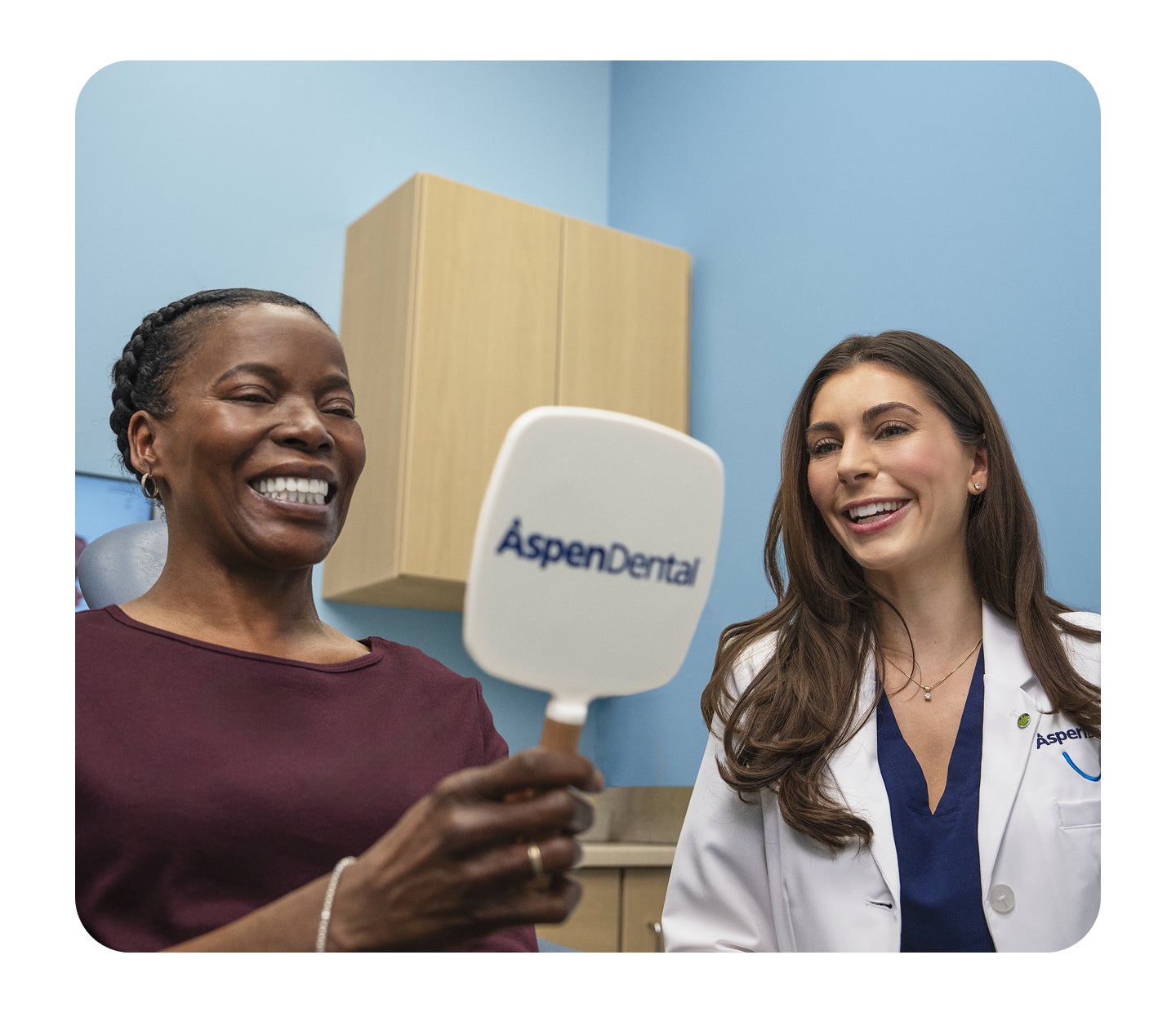 A smiling patient looks into an Aspen Dental mirror while a dentist in a white coat stands beside her, smiling during a dental appointment.