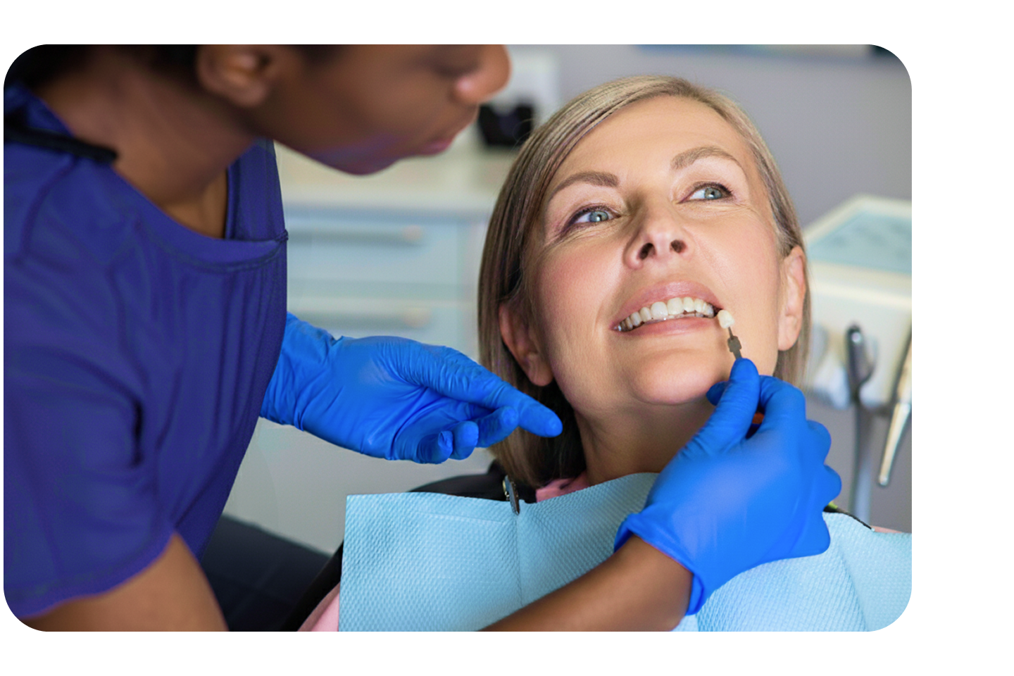 A dental professional wearing blue gloves holds a tooth shade guide next to a smiling female patient’s teeth to match veneer color during a consultation.