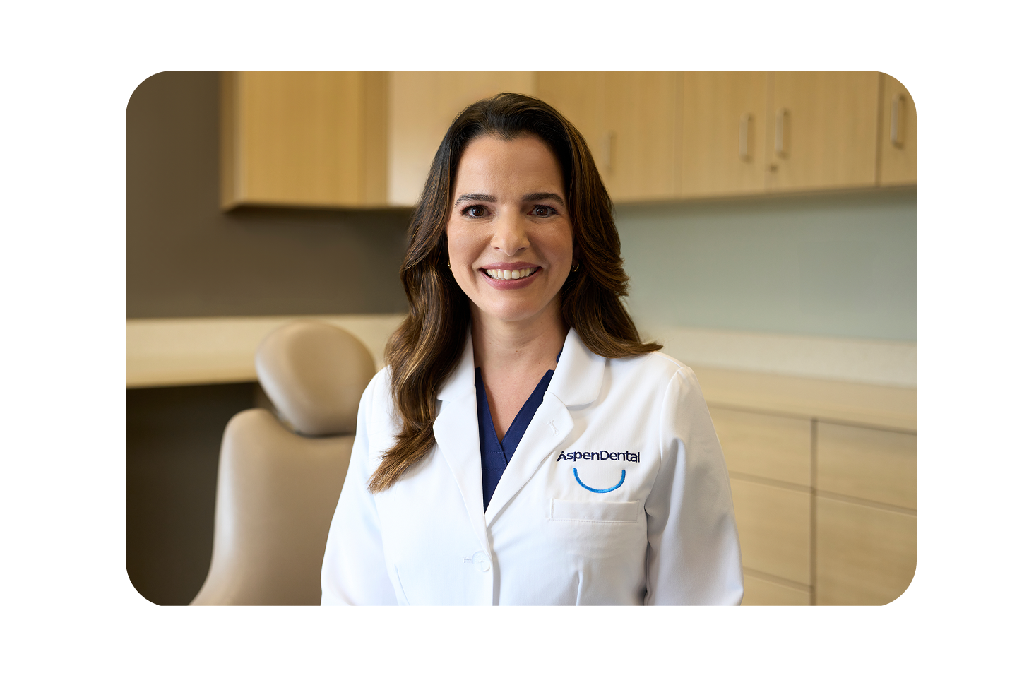 A smiling Aspen Dental provider wearing a white coat stands in a dental exam room with beige cabinets and a patient chair in the background.