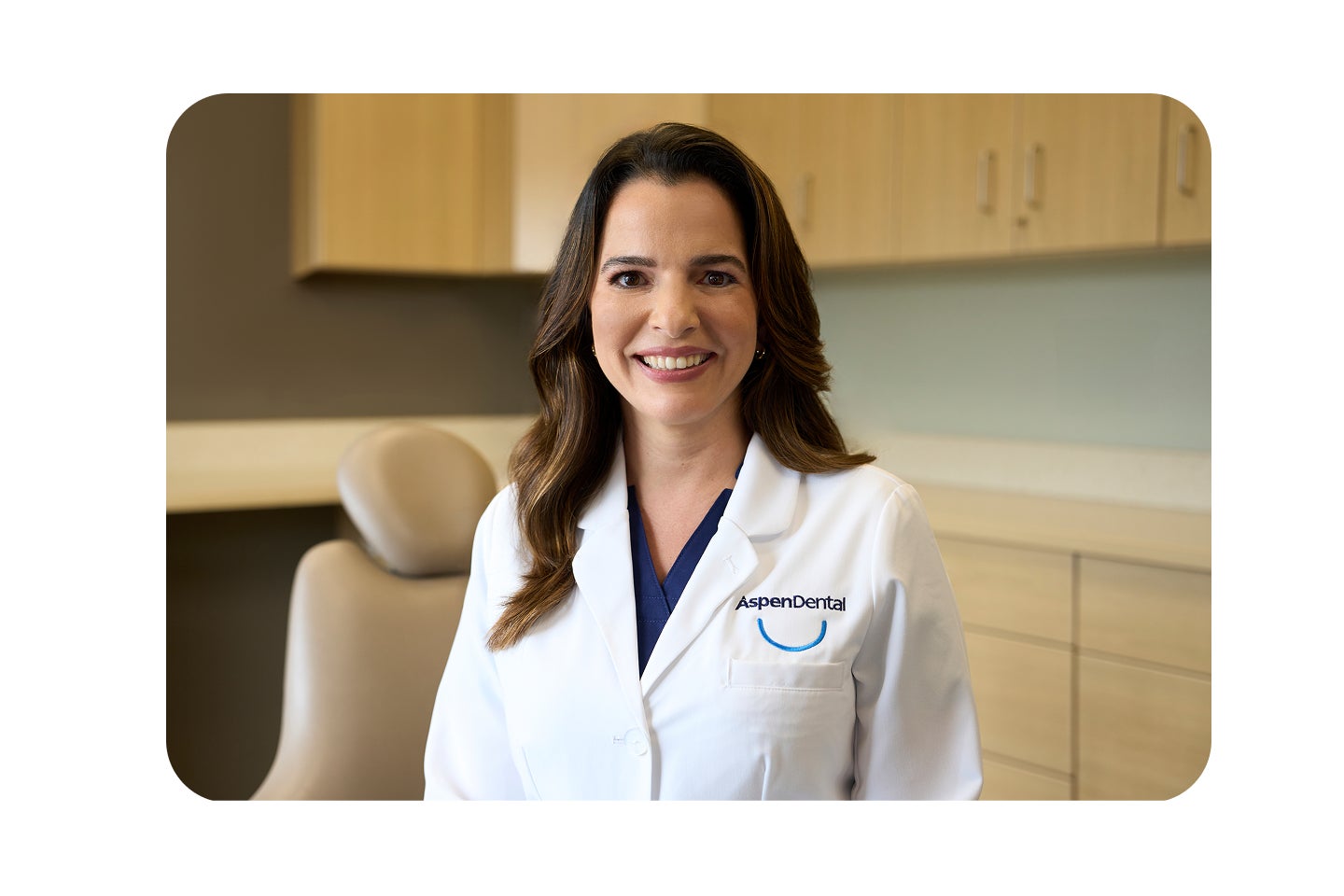 A smiling Aspen Dental provider wearing a white coat stands in a dental exam room with beige cabinets and a patient chair in the background.