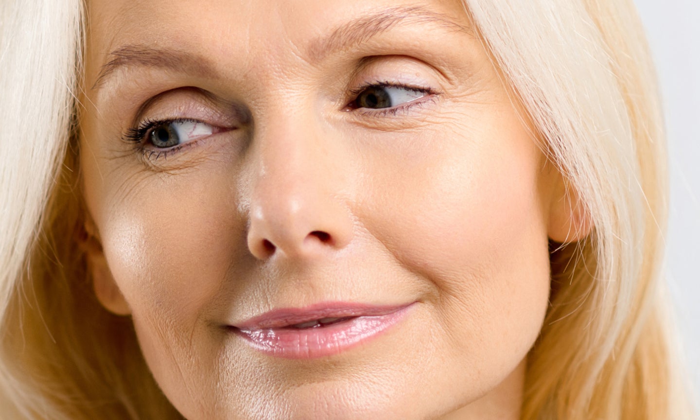 Close-up of a blonde woman with clear, glowing skin and a gentle smile with glossy lips looking slight off to the side