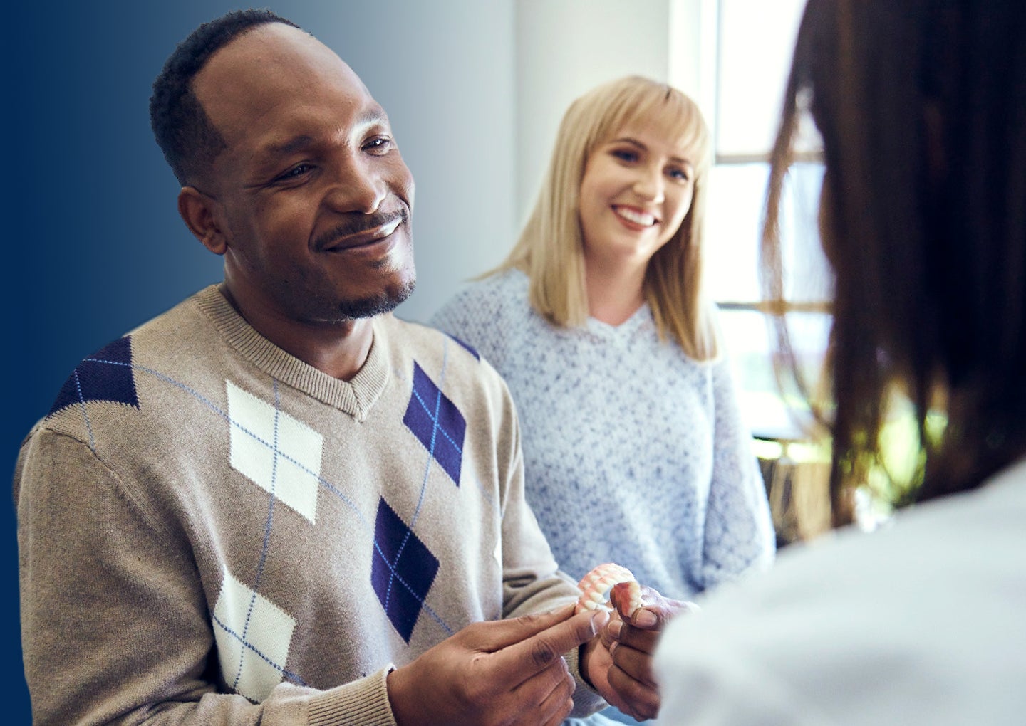 An image of a male patient in a argyle sweater asking a ClearChoice prosthodontist about the dentures trade-in offer during his dental implants consultation, while accompanied by his friend seated next to him.