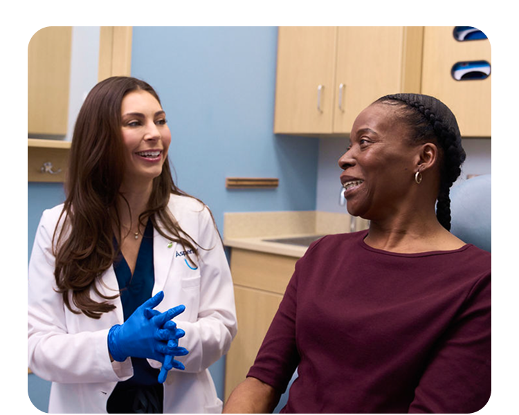 An Aspen Dental doctor in a white coat and blue gloves is smiling while having a discussion with a woman sitting in the dental chair.