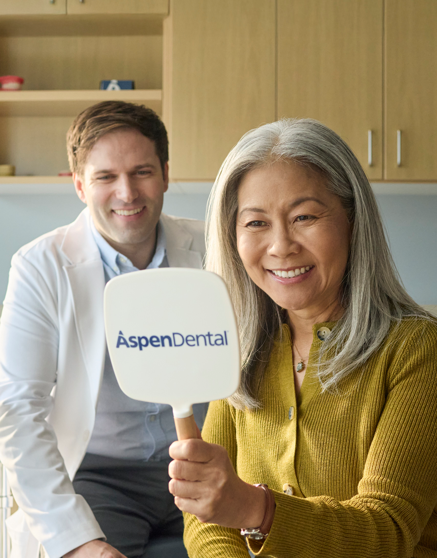 A happy patient admires her smile in an aspen dental mirror, with her dentist in the background