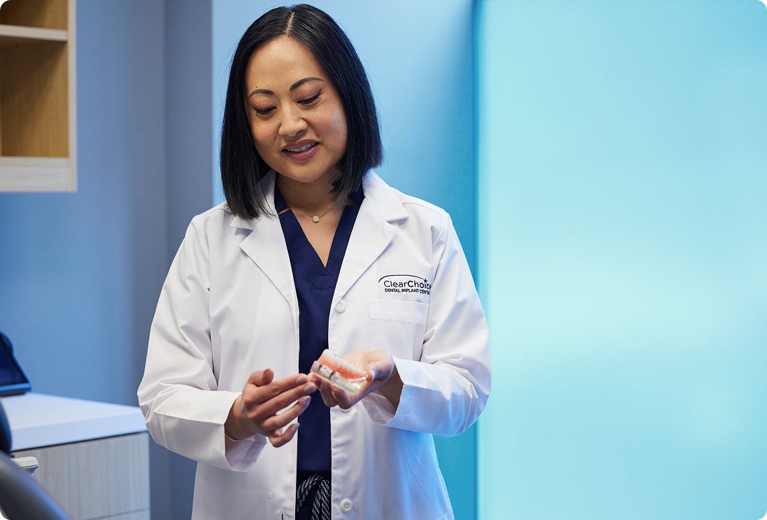 Dr. Theresa Wang, a prosthodontist at ClearChoice, wears a white lab coat and holds a lower arch dental implant model as she explains the components of a dental implant restoration.