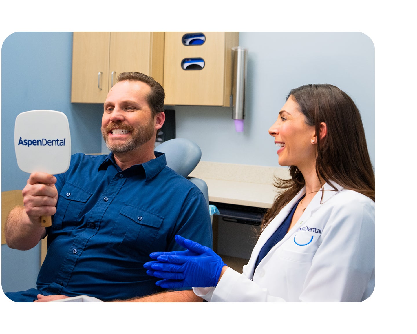 A smiling male patient holds an Aspen Dental mirror to view his teeth while a dentist in a white coat and blue gloves smiles beside him during a dental checkup.