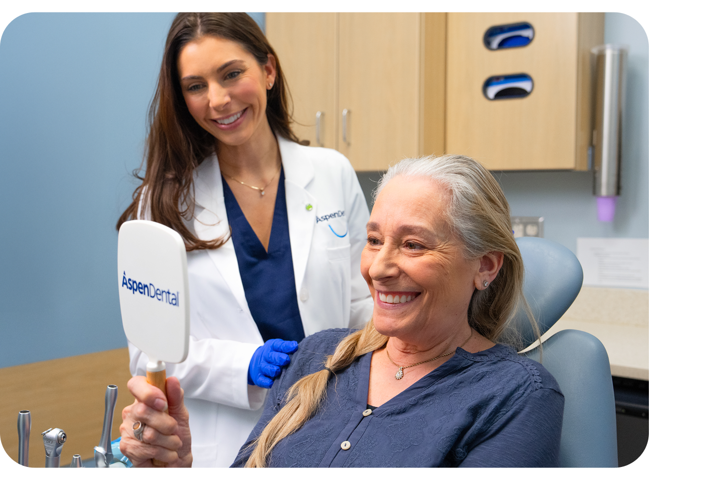 A smiling patient looks into an Aspen Dental mirror while a dentist in a white coat stands beside her during a dental visit.