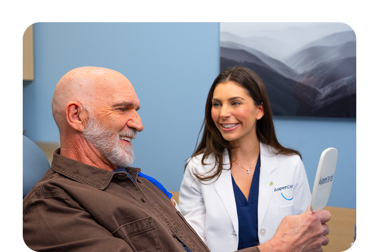 Elderly male patient smiling while holding a mirror as an Aspen Dental dentist smiles beside him during a consultation