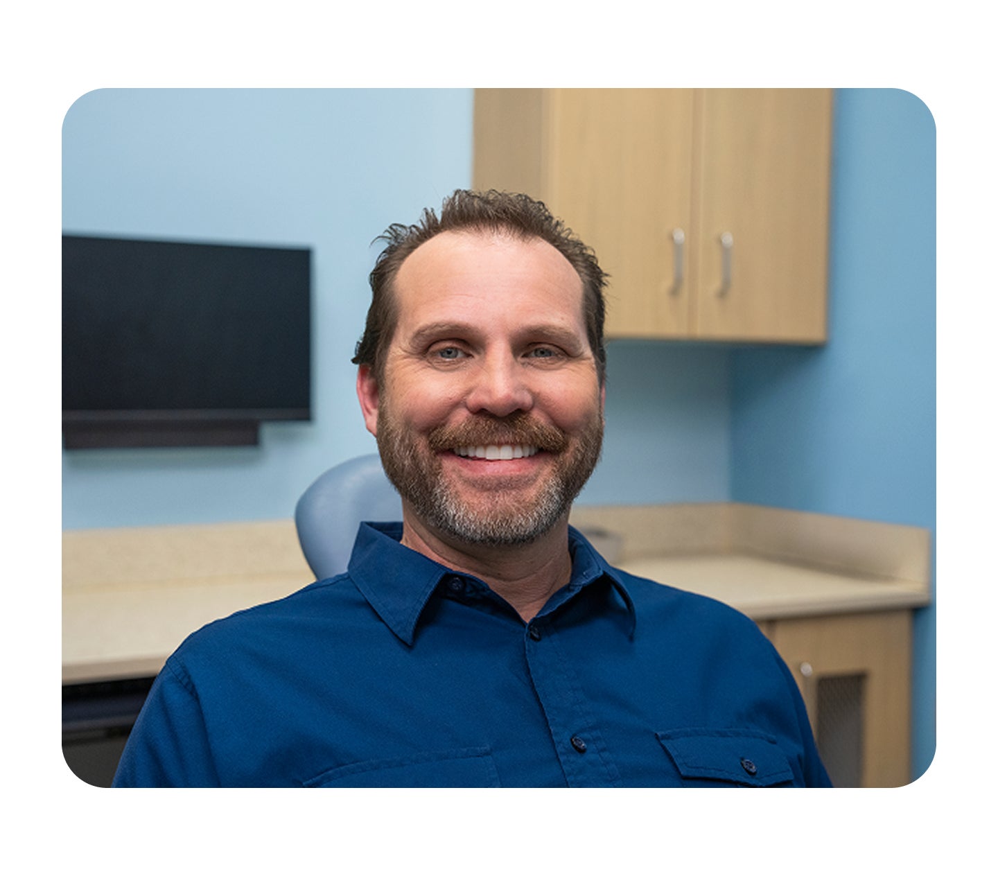 A smiling male patient sits in a dental exam chair wearing a blue shirt, with cabinetry and dental equipment visible in the background of the Aspen Dental office.