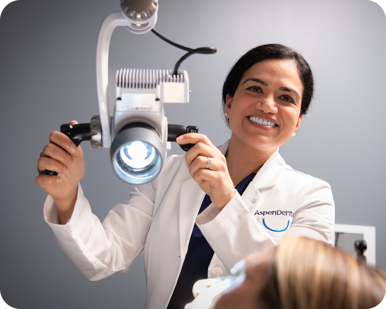 An Aspen Dental dentist in a white coat smiles while adjusting a bright overhead exam light for a patient during a dental appointment.