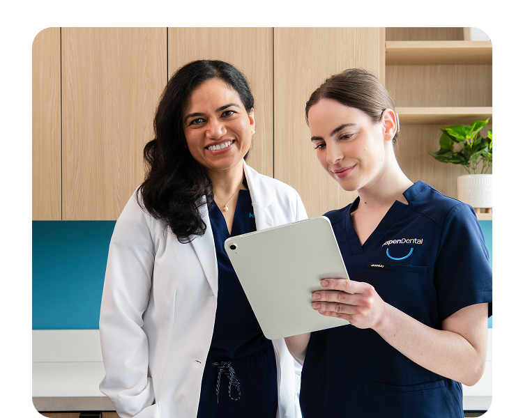 Two Aspen Dental team members, one in a white lab coat and one in scrubs, smiling while reviewing a patient chart on a digital tablet.