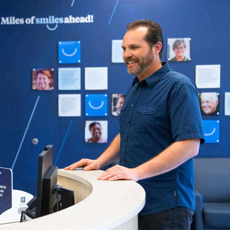 A patient smiles while speaking with an Aspen Dental team member at the front desk, with a blue wall behind them displaying the phrase “Miles of smiles ahead!”.