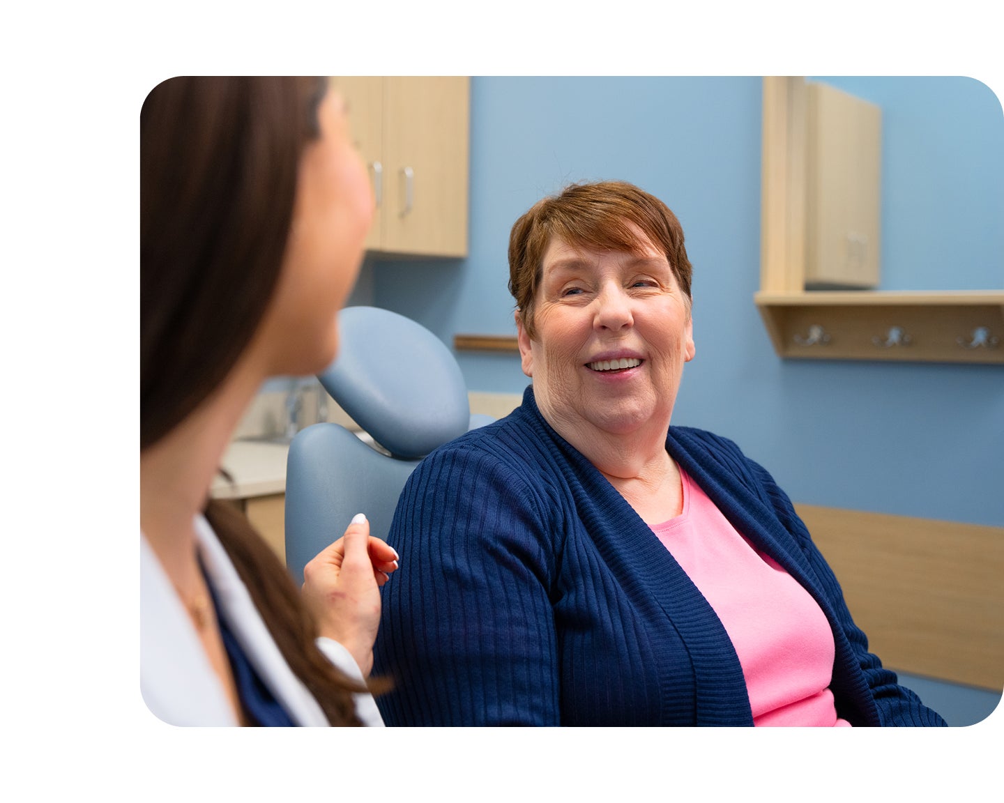 A smiling female patient wearing a pink shirt and navy cardigan talks with her Aspen Dental dentist during a friendly consultation in a dental exam room.