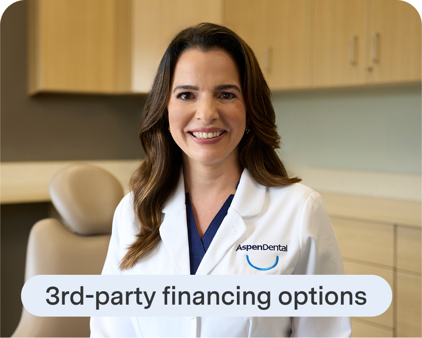 An Aspen Dental dentist in a white coat smiles while standing in a dental exam room with cabinetry and a patient chair in the background.