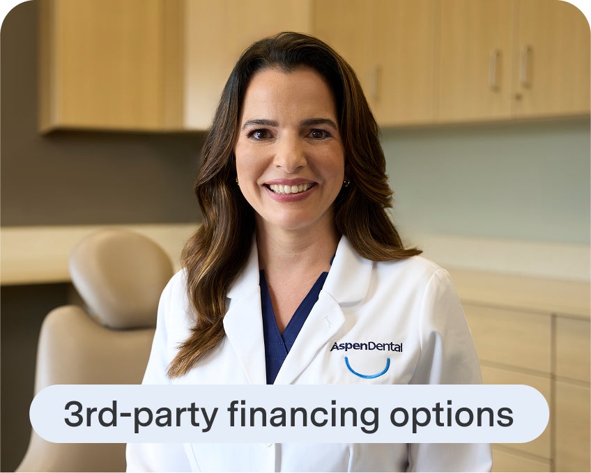 An Aspen Dental dentist in a white coat smiles while standing in a dental exam room with cabinetry and a patient chair in the background.