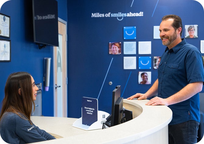 A smiling patient speaks with an Aspen Dental team member at the front desk, with a blue wall behind them displaying the phrase “Miles of smiles ahead!” and patient photos.