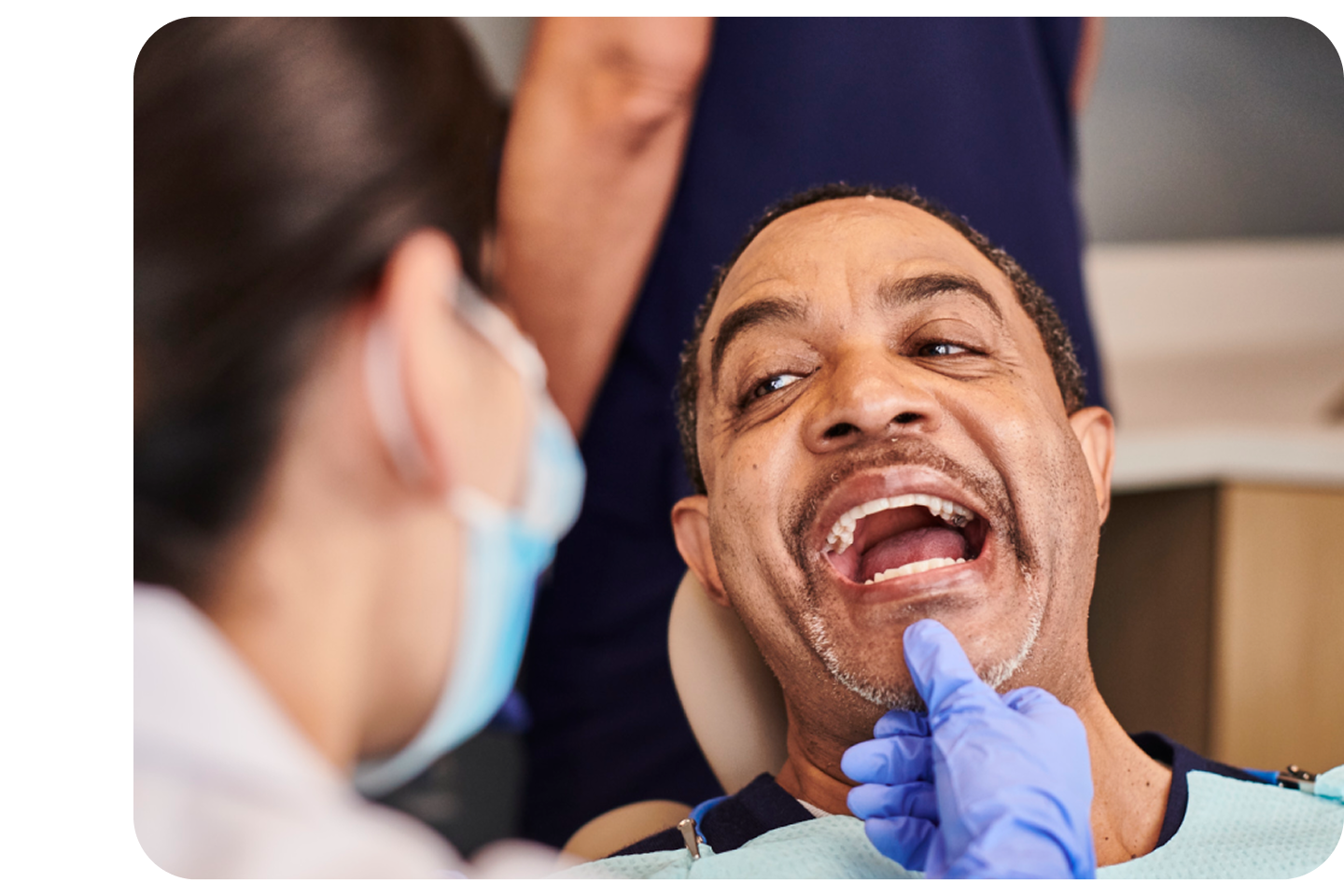 A male patient with a mustache is seated in a dental chair, smiling with his mouth open, as a dentist in a mask and blue glove points to a tooth on his lower jaw during an exam.