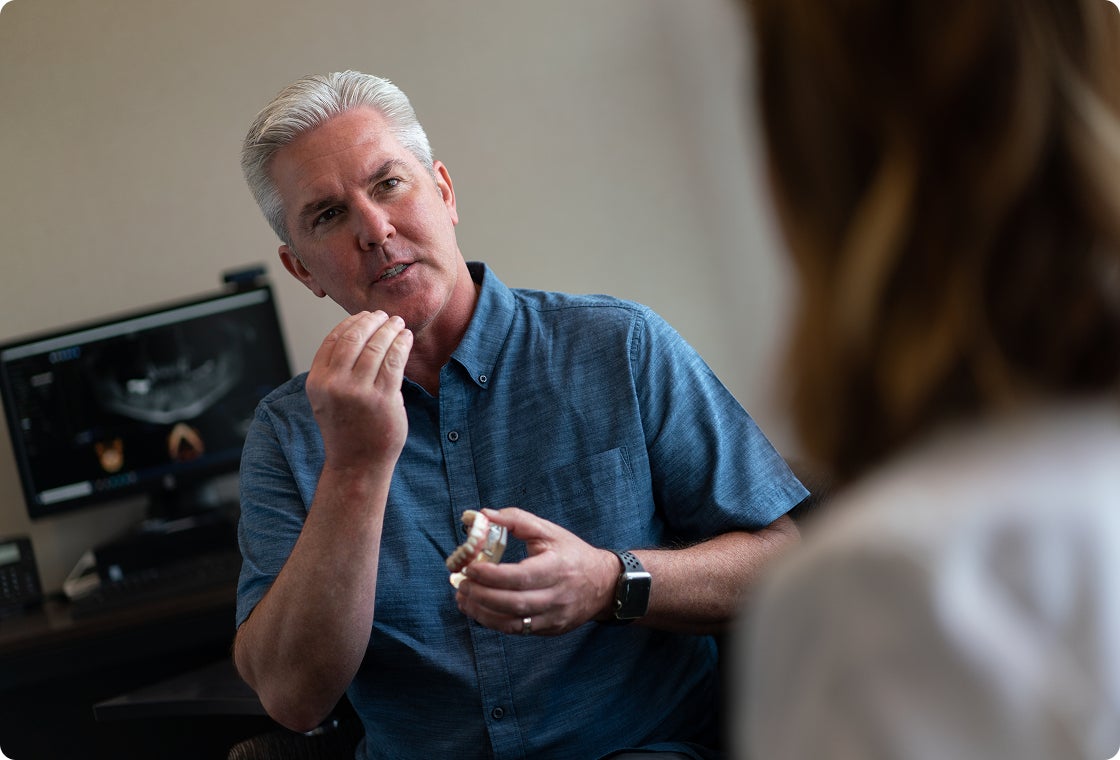 A senior male patient in a blue shirt holds a lower arch teeth model as he asks his prosthodontist questions about his oral health and treatment plan during consultation.