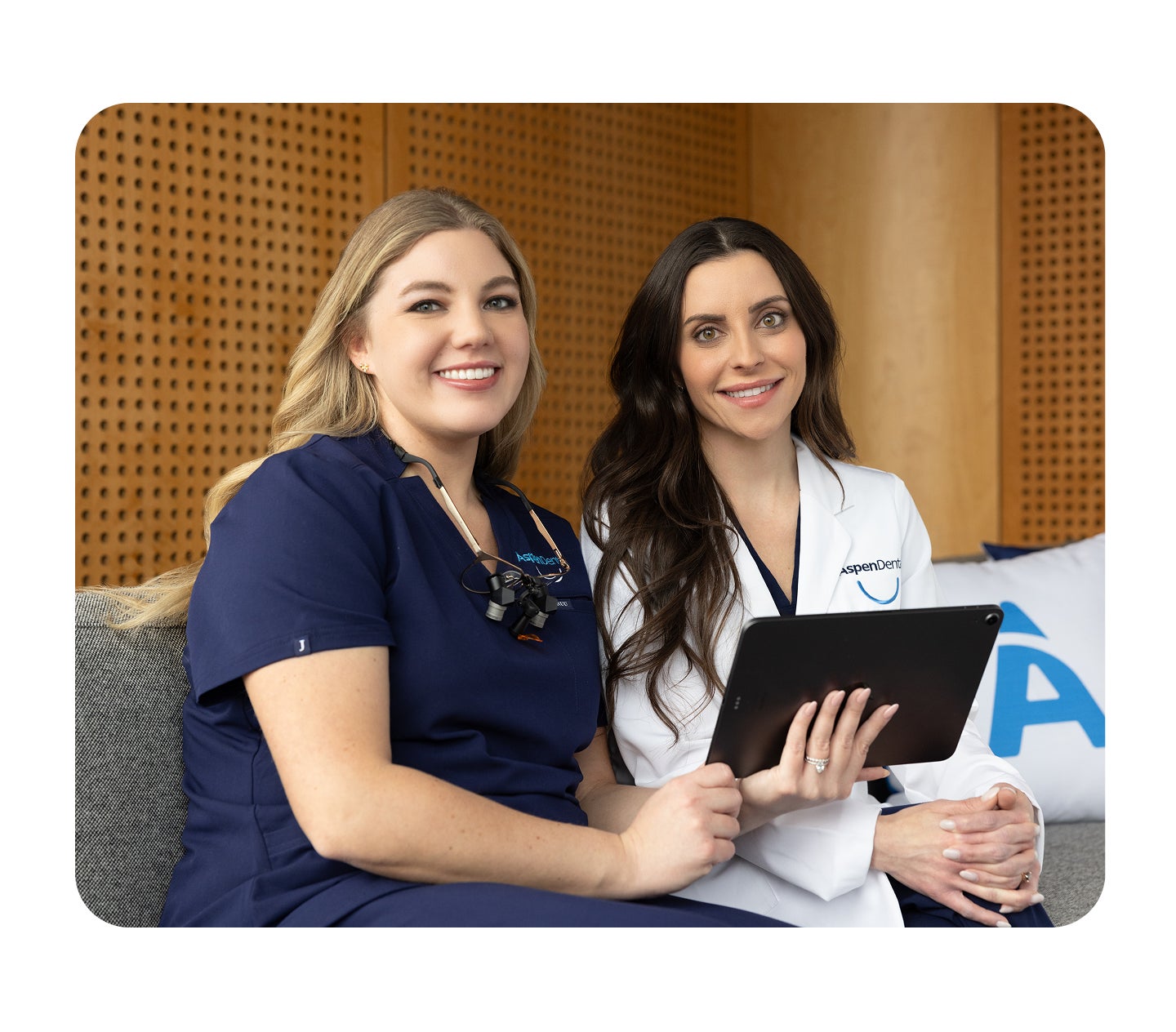 Two Aspen Dental team members, one in navy scrubs and the other in a white coat, smile while sitting together and holding a tablet in a modern office setting.