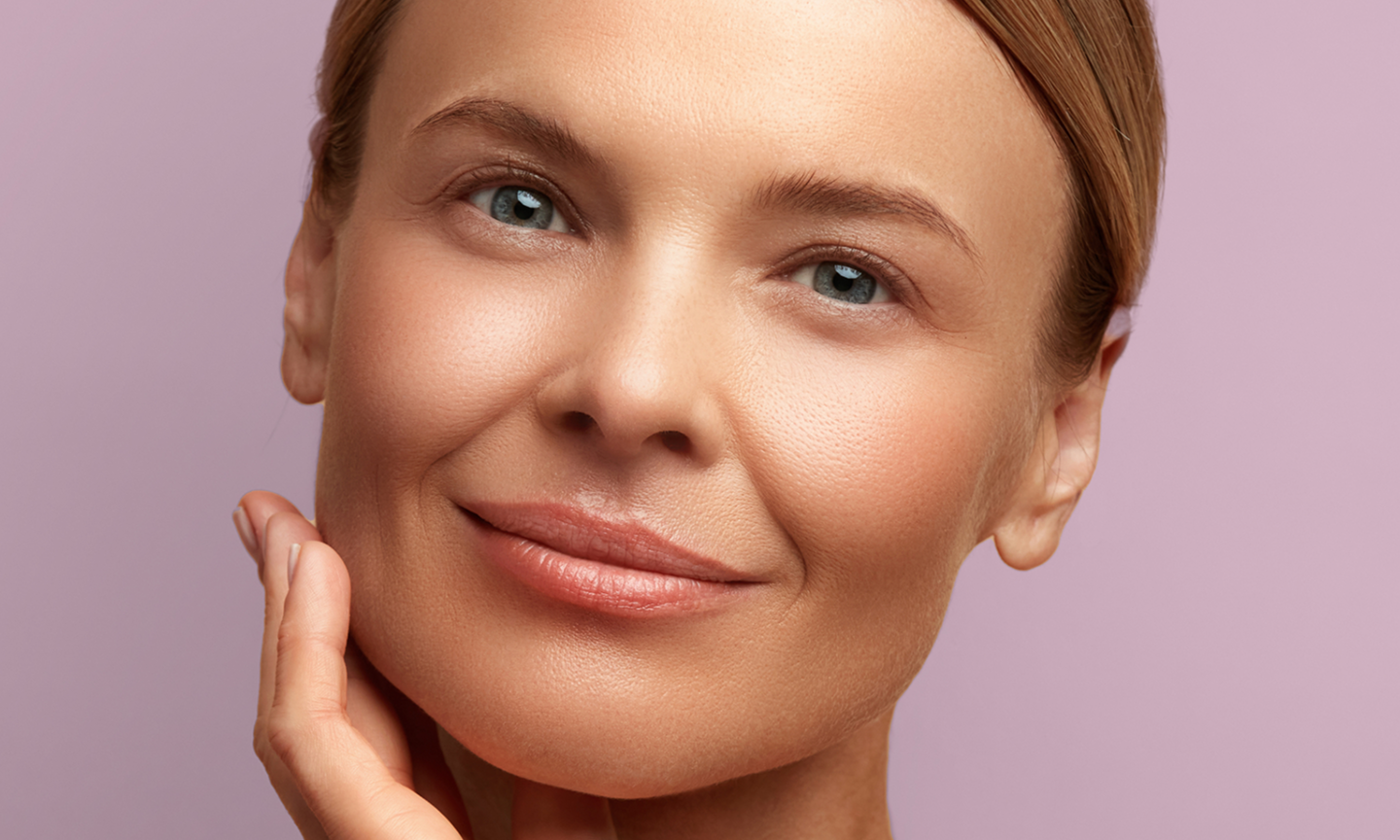 A woman with radiant skin is smiling and touching her face with her hand