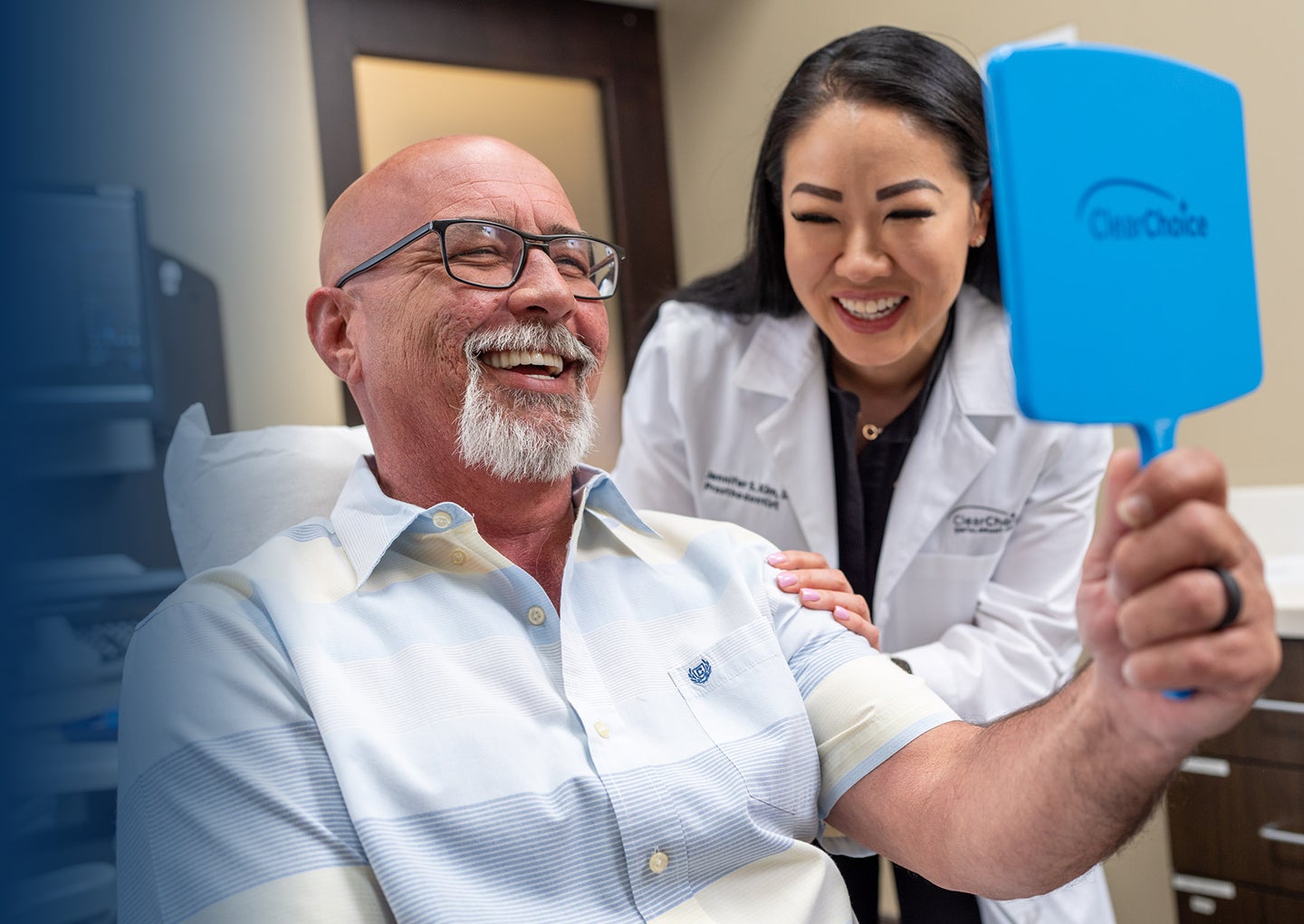 A senior male patient in glasses and a short sleeve shirt smiles into a blue hand held mirror as he celebrates the completion of his dental implant treatment with his prosthodontist standing beside him.
