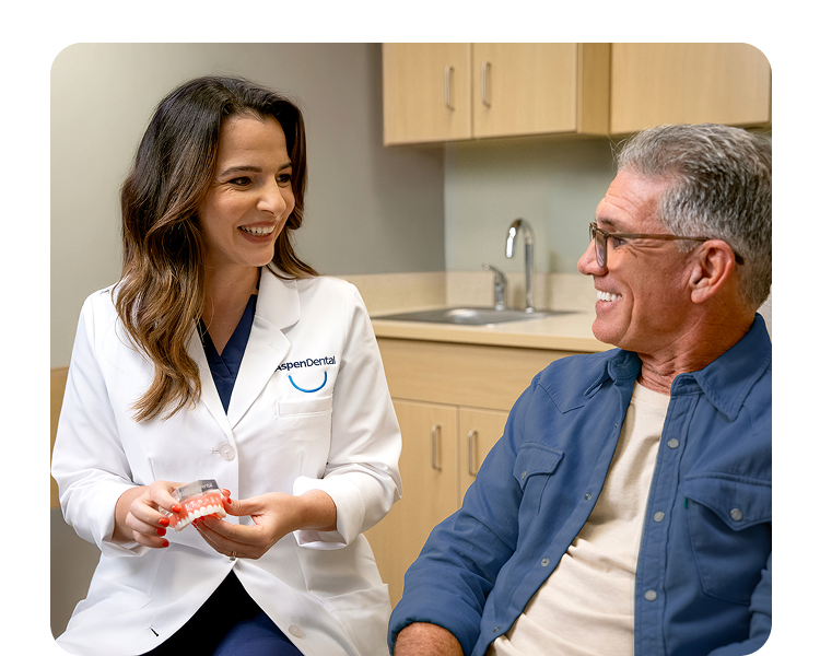 A woman in a dental coat holds a denture while talking to an older man