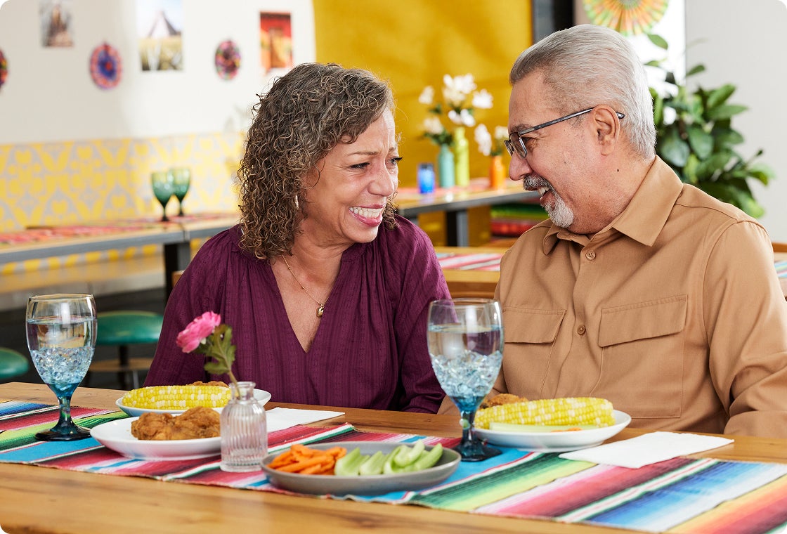 A pair of senior couple smiling at each other as they sit in front of a table filled with food, both overjoyed to be able to enjoy their favorite dishes again after getting dental implants.