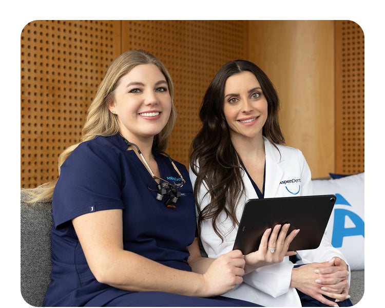 Two Aspen Dental team members, one in navy scrubs and the other in a white coat, smile while sitting together and holding a tablet in a modern office setting.