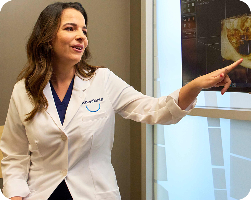 A dental professional in an Aspen Dental lab coat pointing at a screen displaying a dental scan or X-ray