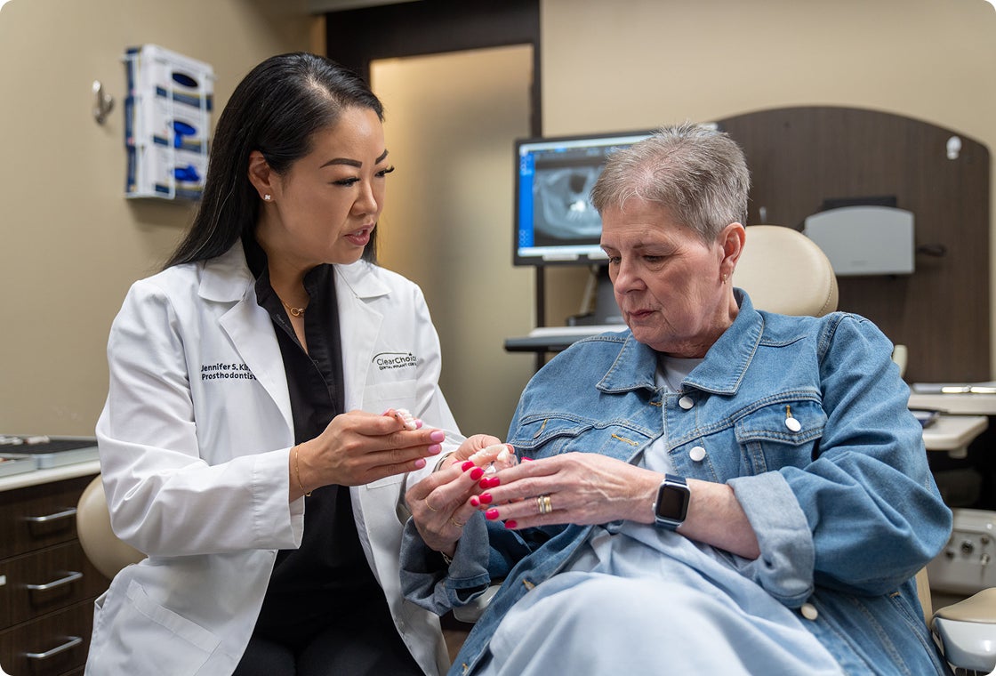 A ClearChoice prosthodontist in a white lab coat shows an upper and lower arch dental implant model to a senior patient sitting in a dental chair, while explaining to her the treatment plan and timeline.