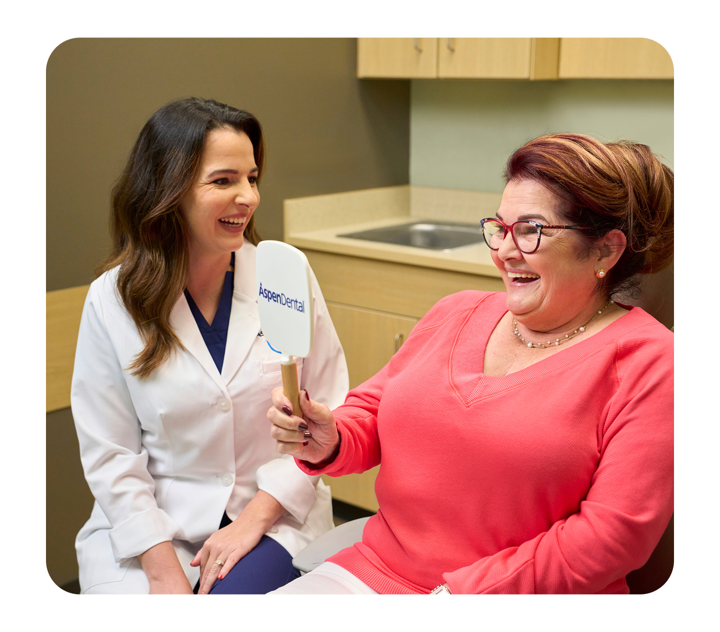 A happy patient wearing glasses smiles while looking into an Aspen Dental mirror, as a dentist in a white coat sits beside her and smiles during the appointment.