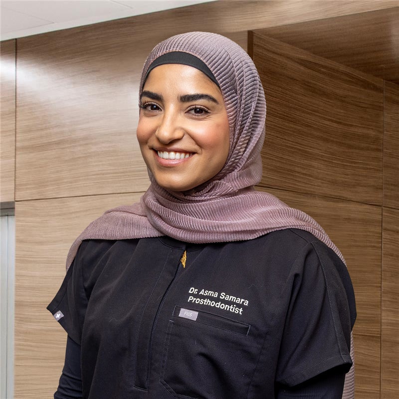 Dr. Asma Samara smiling in front of a brown wood office wall