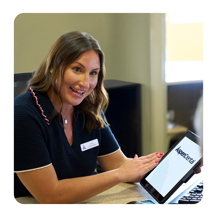 An Aspen Dental team member smiles while explaining information on a tablet to a patient during a consultation at the front desk.