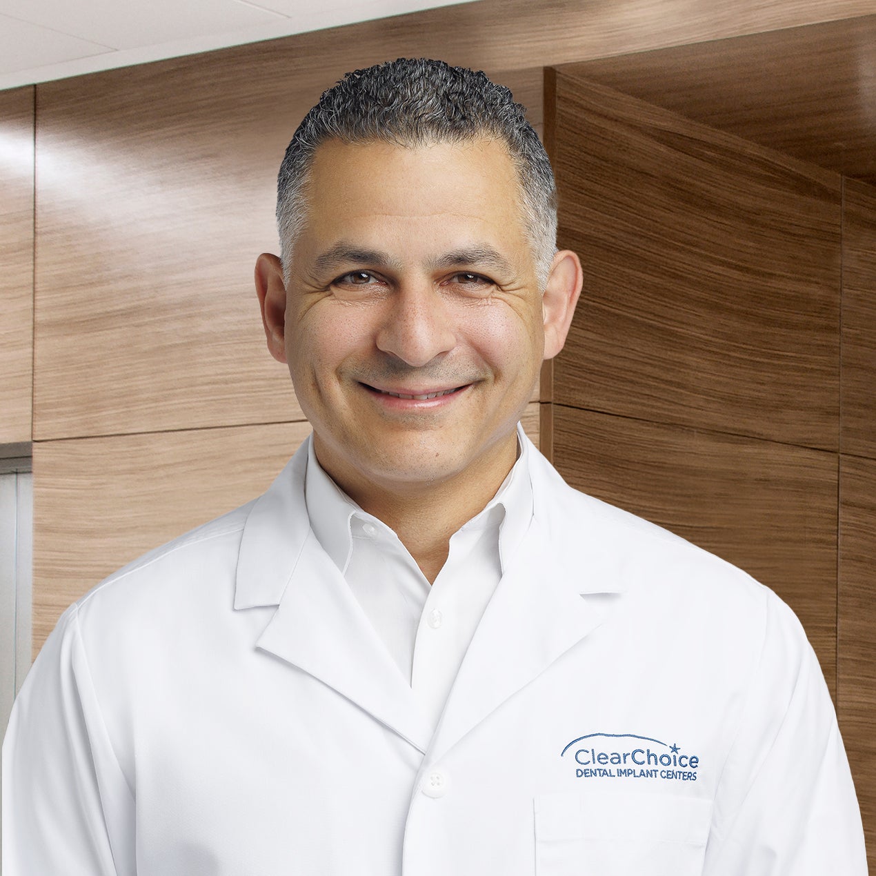 Dr. Mohamed Kamel smiling in a white ClearChoice coat in front of a brown wood office wall