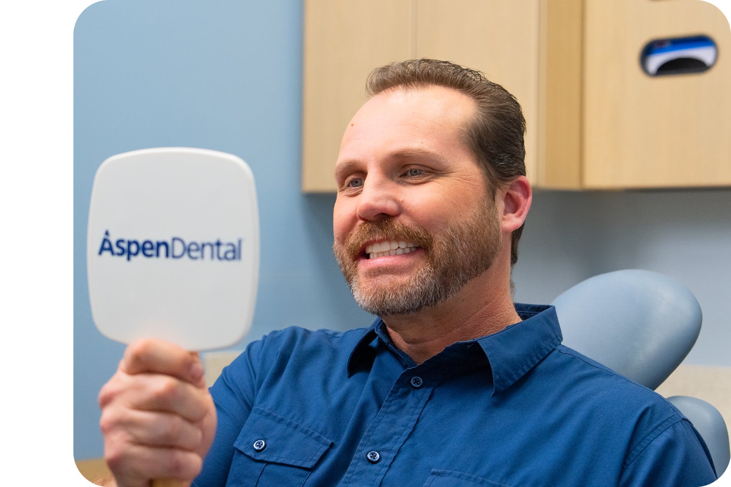 A male patient sitting in a dental exam chair smiles while looking at his reflection in an Aspen Dental hand mirror.