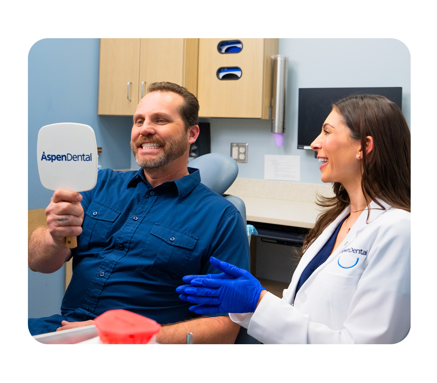 An Aspen Dental provider smiles while sitting next to a patient in a dental chair who is holding up an Aspen Dental-branded handheld mirror in a dental office setting