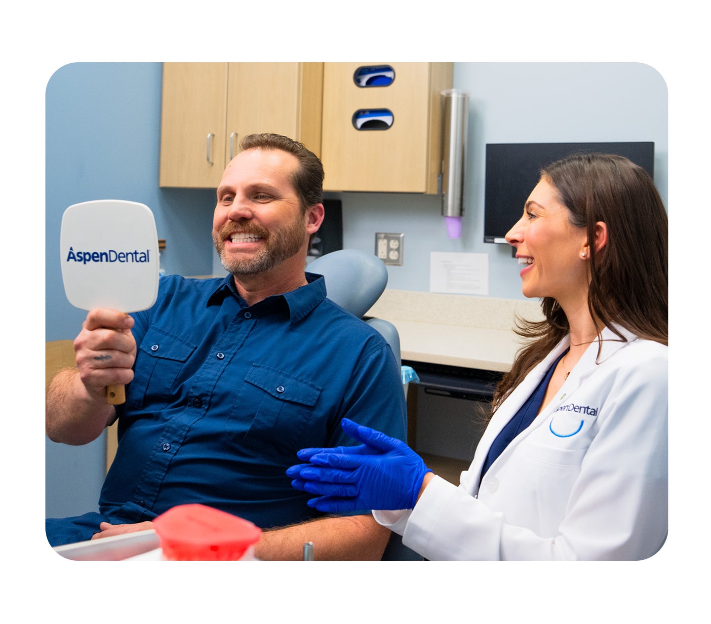 An Aspen Dental provider smiles while sitting next to a patient in a dental chair who is holding up an Aspen Dental-branded handheld mirror in a dental office setting