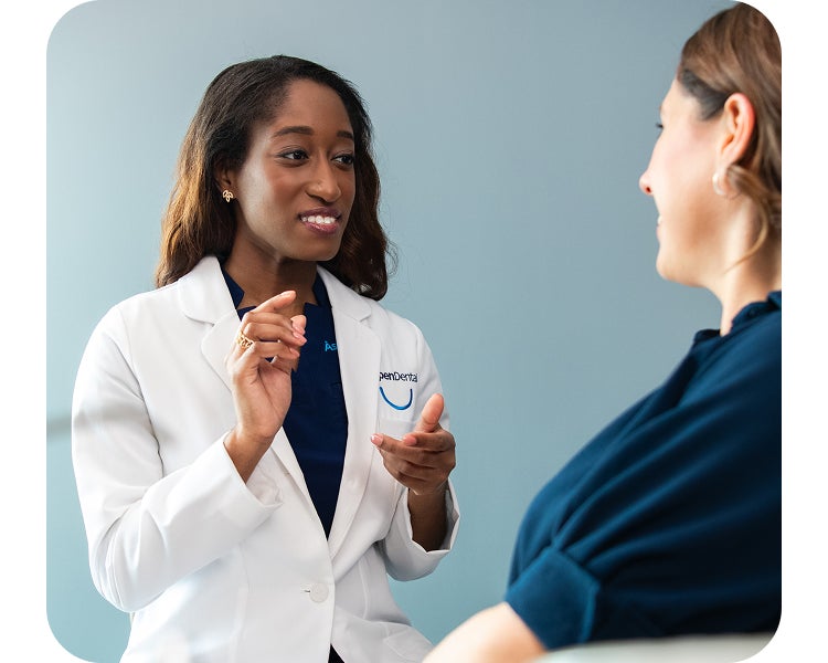 An Aspen Dental dentist in a white coat speaks with a seated patient, offering guidance and answering questions during a consultation in a modern exam room.