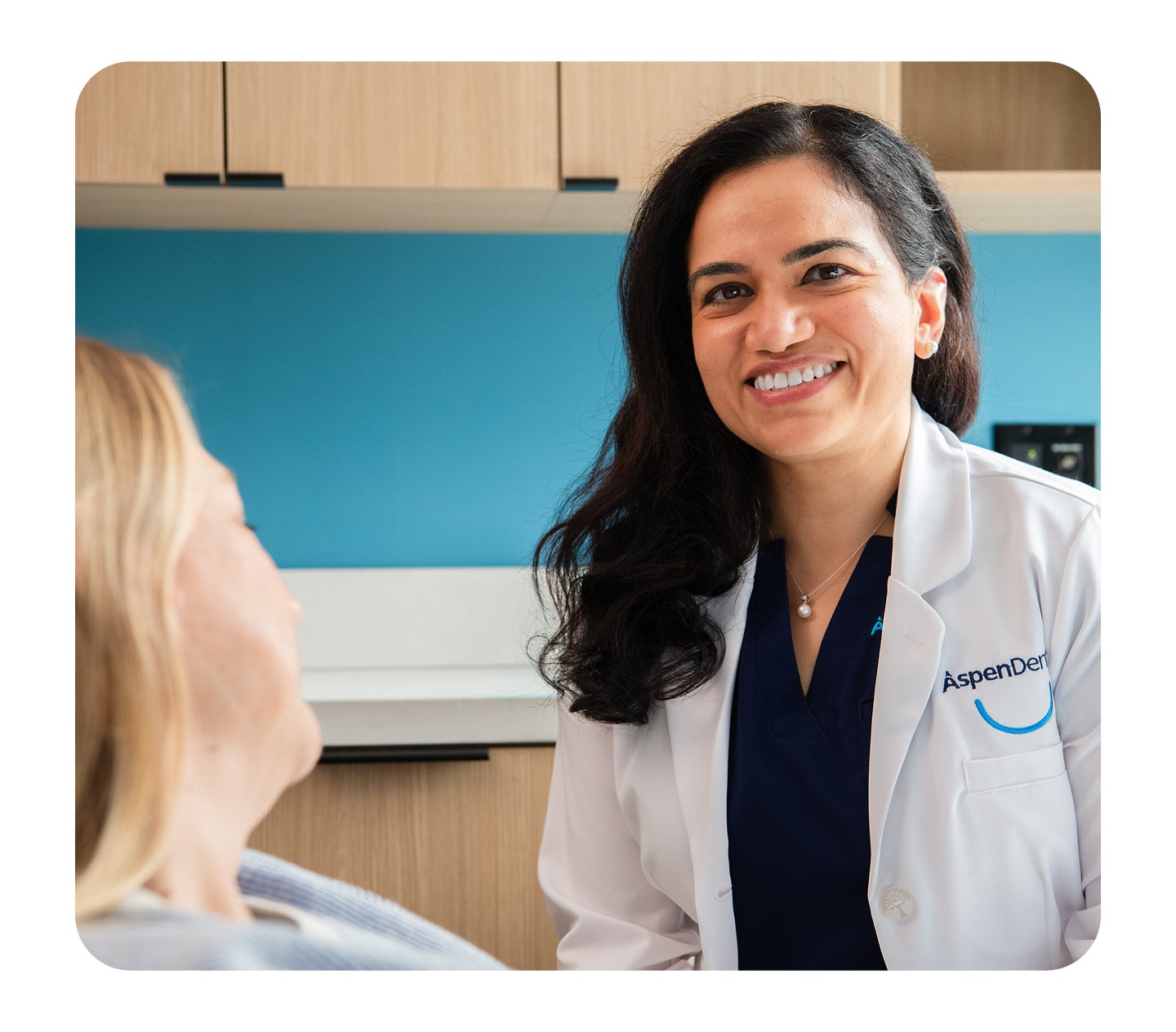 Aspen Dental dentist in a white coat smiling confidently at a female patient during a consultation in an exam room.