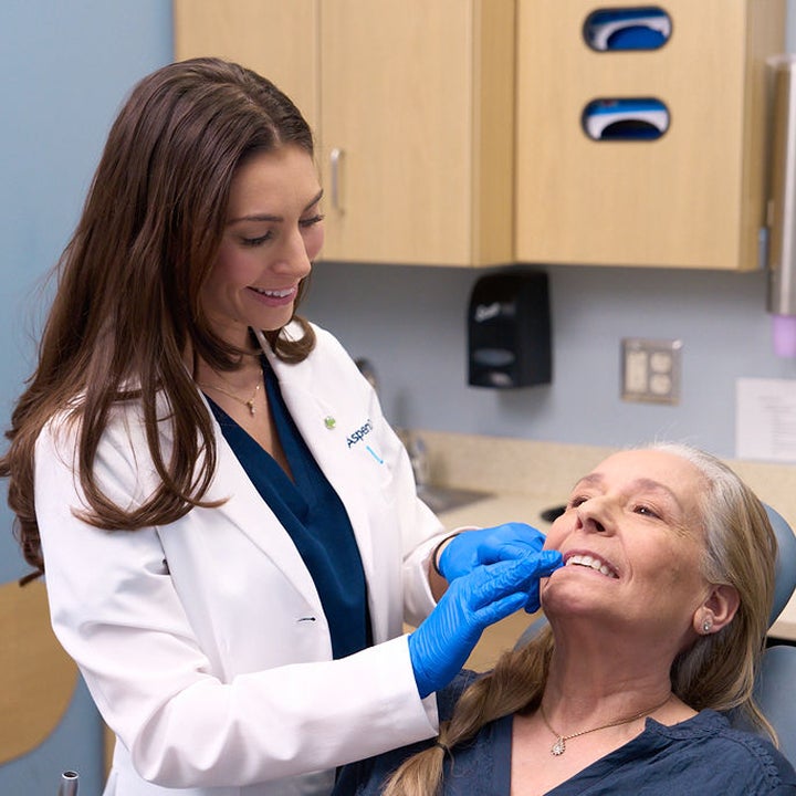 An Aspen Dental dentist wearing blue gloves fits a dental appliance or aligner for a smiling female patient seated in a dental exam chair.