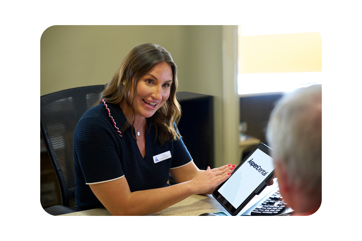 An Aspen Dental team member smiles while showing information on a tablet to a patient at the front desk.