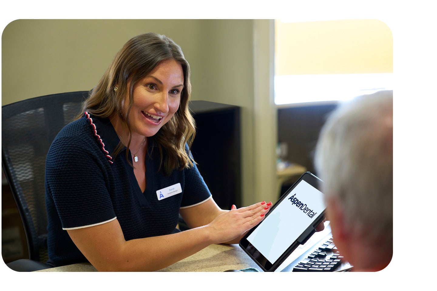 A friendly Aspen Dental team member smiles while showing a patient information on a tablet displaying the Aspen Dental logo during a financial consultation.