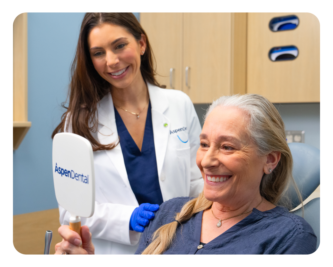 A happy patient admires her smile in an aspen dental mirror, with her dental professional looking on