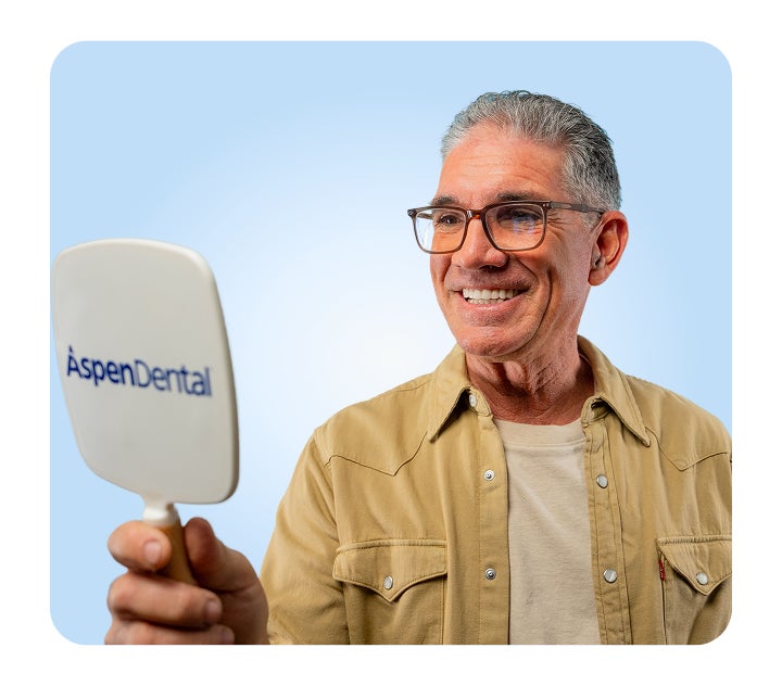A smiling male patient wearing glasses admires his reflection in an Aspen Dental mirror against a light blue background.