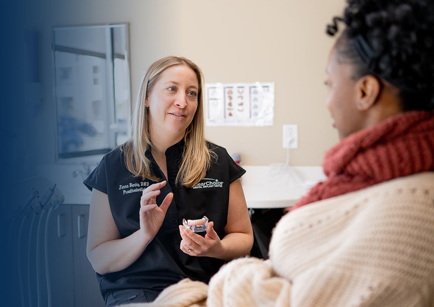 A ClearChoice provider wearing a navy blue scrub uses a lower arch dental implant model to explain the implant process to a patient, who is wearing a red scarf and white sweater while sitting in a dental chair, during an in-office consultation.