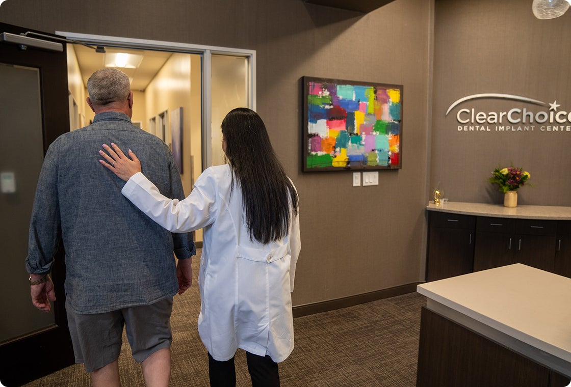 A ClearChoice prosthodontist in a white lab coat pats a male patient on the back as she provides him with support and reassurance before his dental implant consultation.