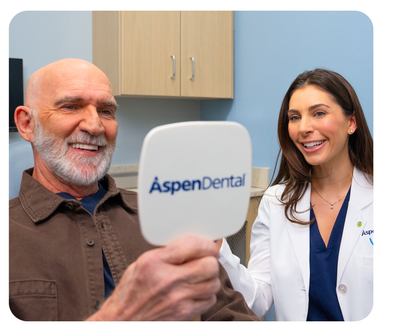 A happy Aspen Dental patient admires his smile in a handheld mirror after receiving dental implants as a smiling Aspen Dental dentist stands next to him with her hand on the patients shoulder. The image conveys confidence symbolizing the benefits of dental implants.