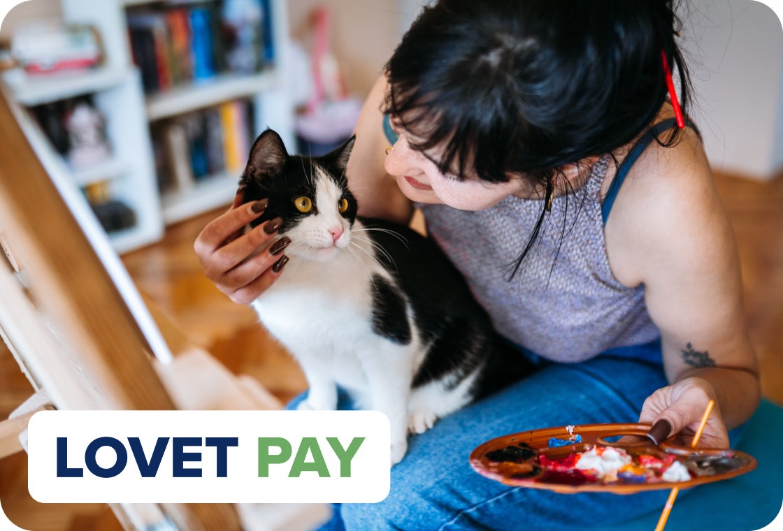 Woman sitting on the floor painting, holding a black-and-white cat beside an easel, with a paint palette in her hand and a “Lovet Pay” logo displayed in the corner.