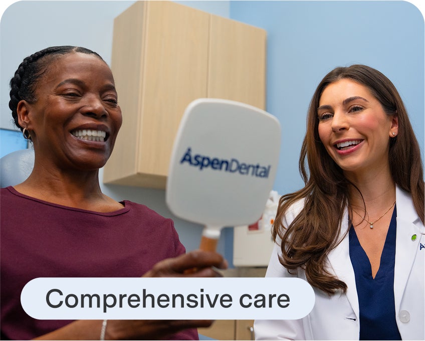 A smiling female dentist in a white lab coat sitting next to a happy female patient who is looking at her teeth in a mirror with the Aspen Dental logo on the back. The text "Comprehensive care" is overlaid on the image.